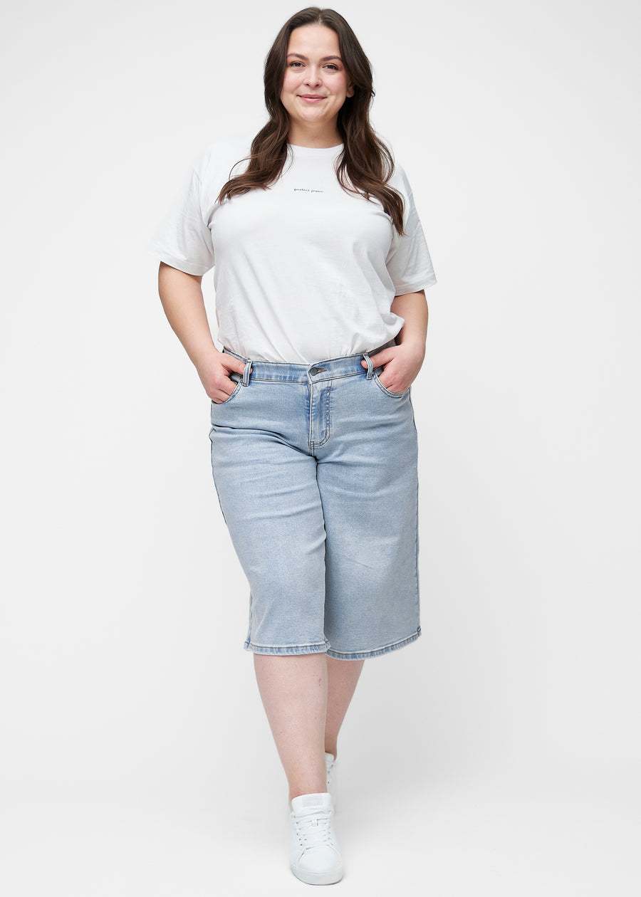 Fuldt billede af plus-size model i løse, lyseblå denim capris i farven Waves, der går til lige under knæet.