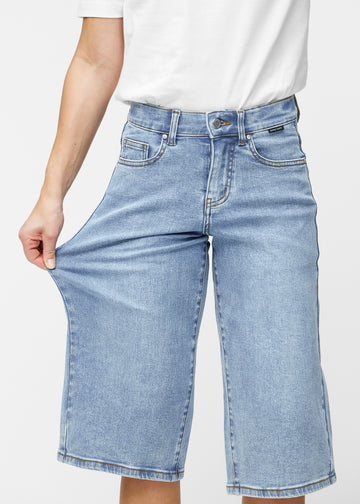 Model strækker de løse, lyseblå denim capris i farven Waves ved hoften for at vise materialets stretch.