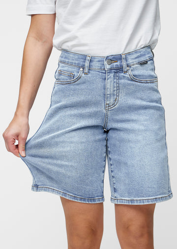 Model strækker de løse, lyseblå denim shorts i farven Waves ved hoften for at vise materialets stretch.