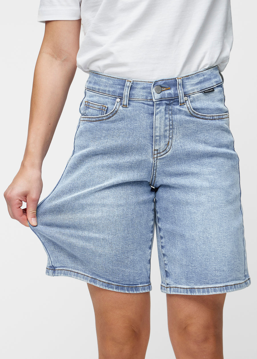 Model strækker de løse, lyseblå denim shorts i farven Waves ved hoften for at vise materialets stretch.