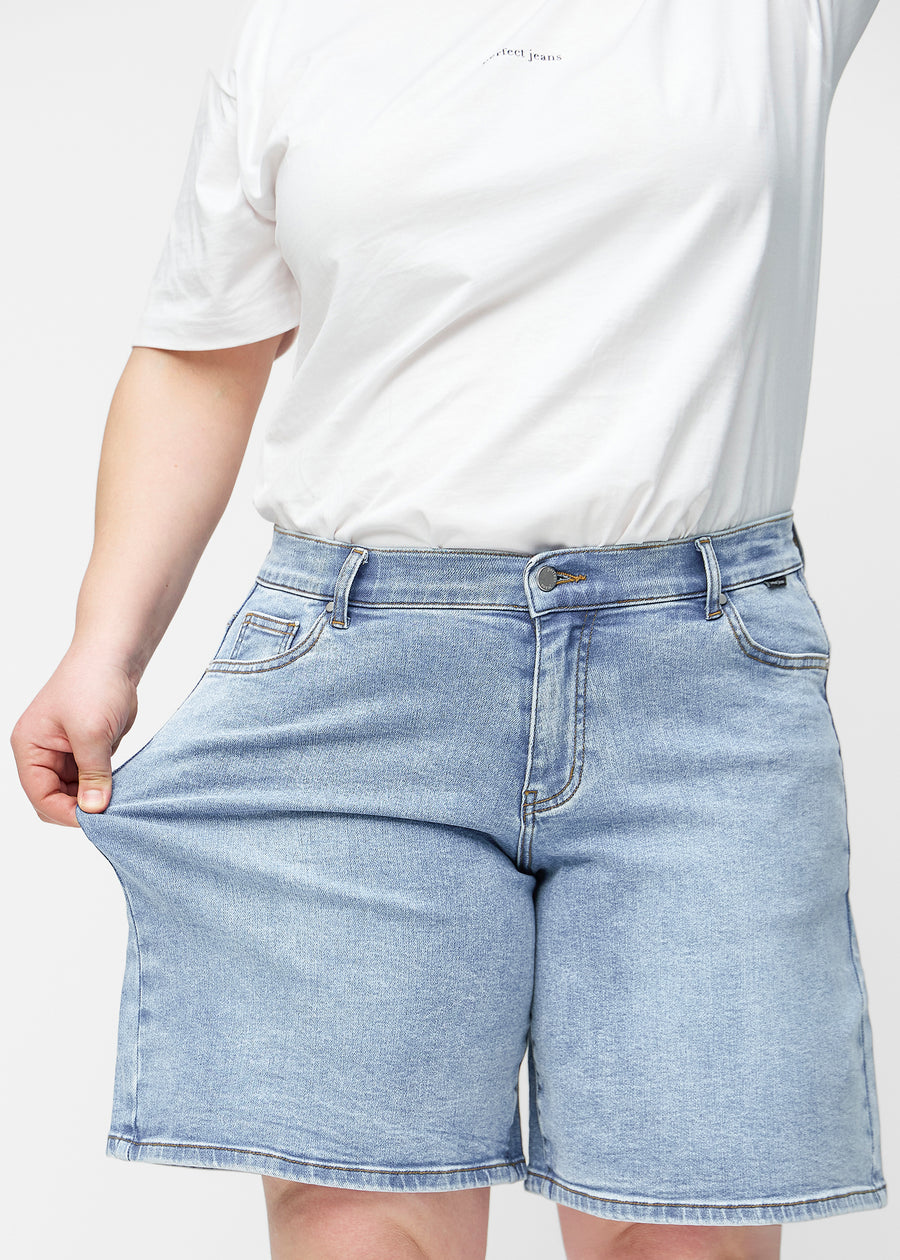 Plus-size model strækker de løse, lyseblå denim shorts i farven Waves ved hoften for at vise materialets stretch.