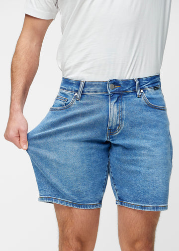 Model strækker blå denim shorts til mænd i farven Rivers ved låret for at vise materialets stretch.