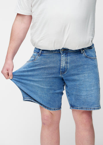 Plus-size model strækker blå denim shorts til mænd i farven Rivers ved låret for at vise materialets stretch.