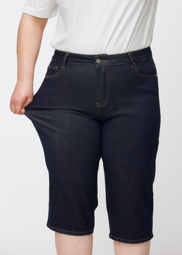 Plus-size model strækker dyb mørkeblå denim capris til kvinder i farven Midnights ved låret for at vise materialets stretch.