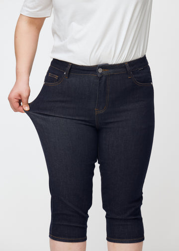 Plus-size model strækker dyb mørkeblå denim capris til kvinder i farven Midnights ved låret for at vise materialets stretch.