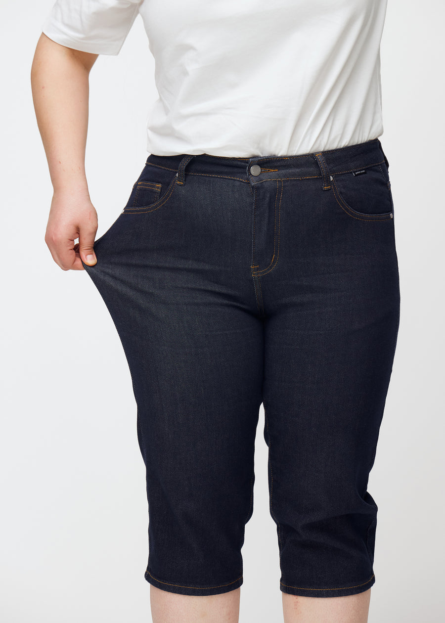 Plus-size model strækker dyb mørkeblå denim capris til kvinder i farven Midnights ved låret for at vise materialets stretch.