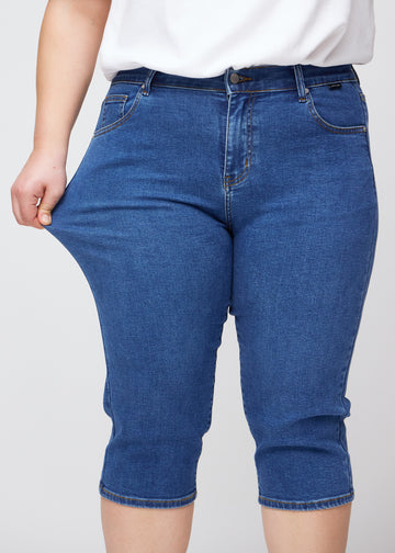 Plus-size model viser stretch ved at trække i caprisene ved låret – fokus på komfort og fleksibilitet i den mørkeblå denim.