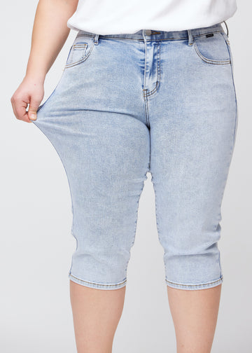 Plus-size model viser stretch ved at trække i caprisene ved låret – fokus på komfort og fleksibilitet i den lyseblå denim.