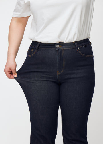 Plus-size model strækker dyb mørkeblå denim jeans til kvinder i farven Midnights ved låret for at vise materialets stretch.