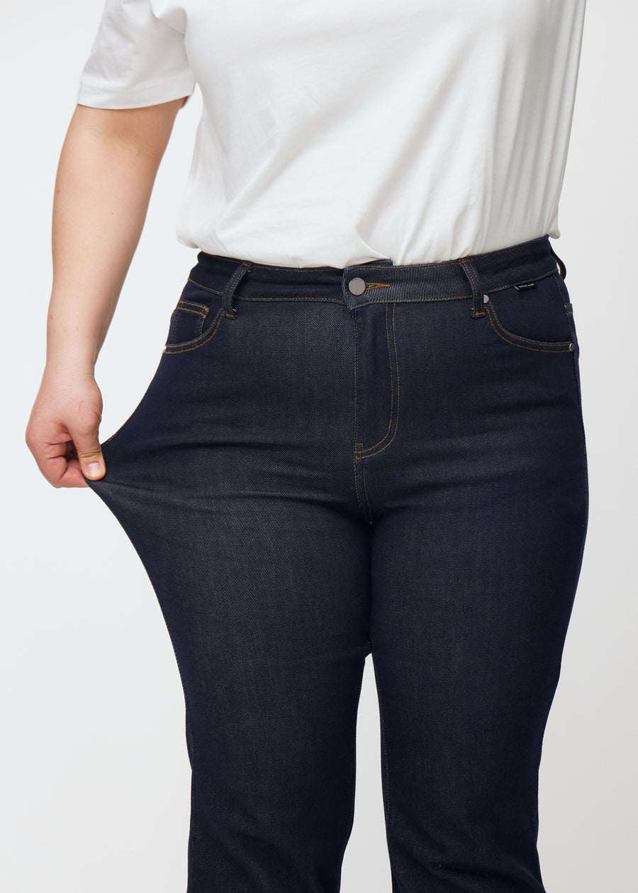 Plus-size model strækker dyb mørkeblå denim jeans til kvinder i farven Midnights ved låret for at vise materialets stretch.