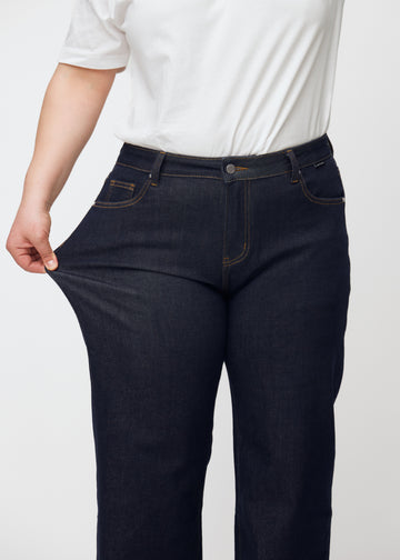 Plus-size model strækker dyb mørkeblå denim jeans til kvinder i farven Midnights ved låret for at vise materialets stretch.