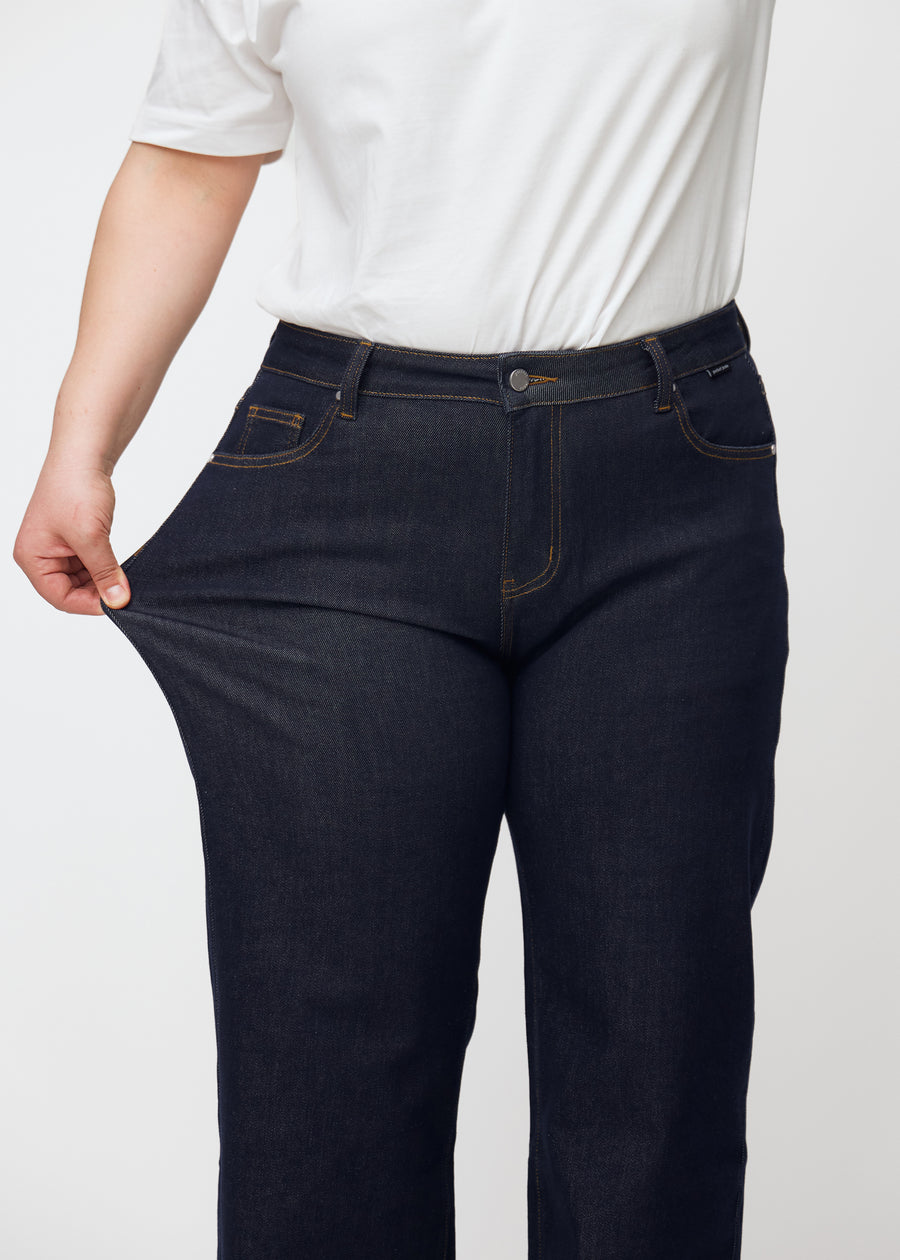 Plus-size model strækker dyb mørkeblå denim jeans til kvinder i farven Midnights ved låret for at vise materialets stretch.