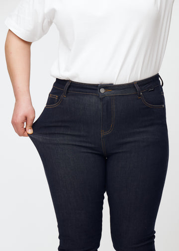 Plus-size model strækker dyb mørkeblå denim jeans til kvinder i farven Midnights ved låret for at vise materialets stretch.