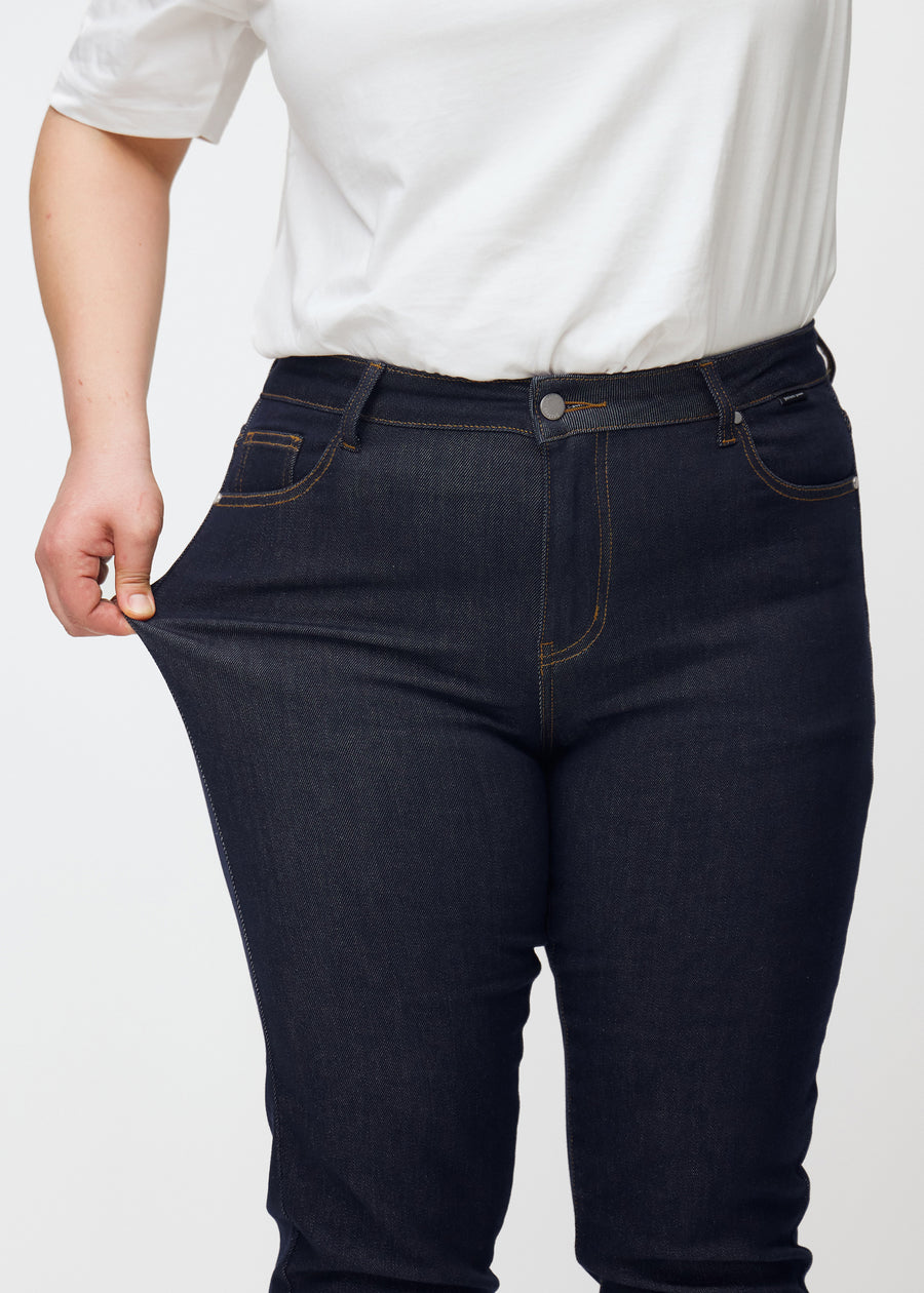 Plus-size model strækker dyb mørkeblå denim jeans til kvinder i farven Midnights ved låret for at vise materialets stretch.