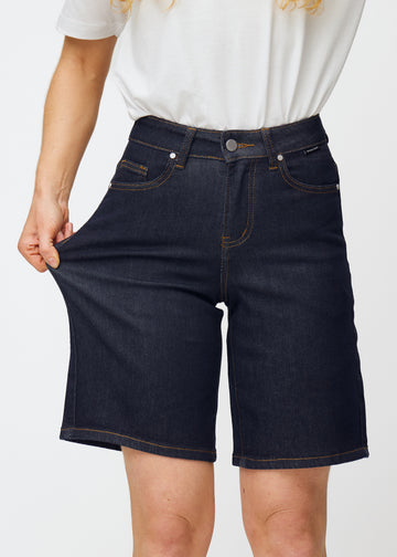 Model strækker dyb mørkeblå denim shorts til kvinder i farven Midnights ved låret for at vise materialets stretch.