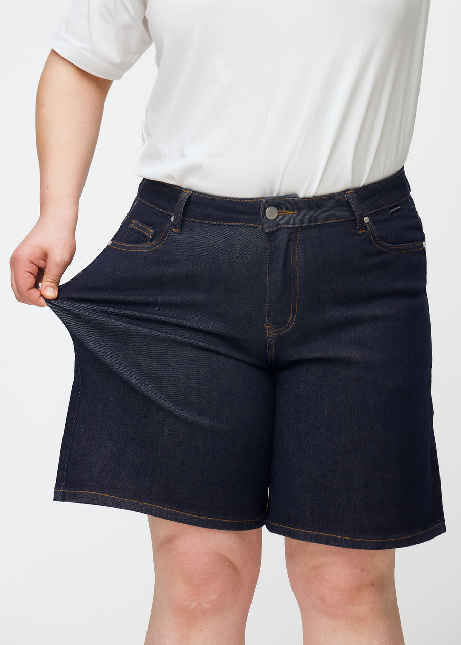 Plus-size model strækker dyb mørkeblå denim shorts til kvinder i farven Midnights ved låret for at vise materialets stretch.