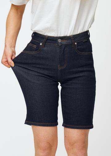 Model strækker dyb mørkeblå denim shorts til kvinder i farven Midnights ved låret for at vise materialets stretch.