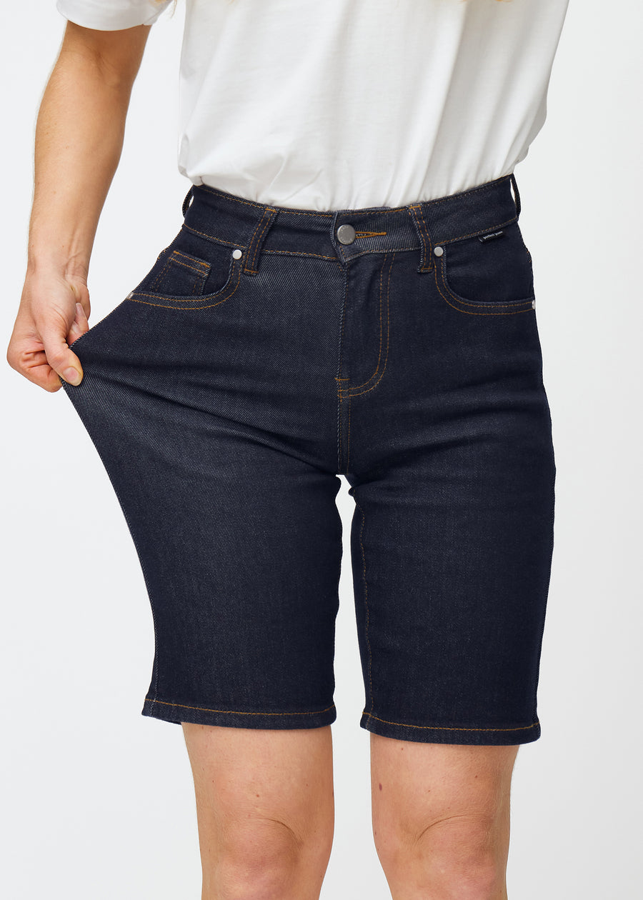 Model strækker dyb mørkeblå denim shorts til kvinder i farven Midnights ved låret for at vise materialets stretch.