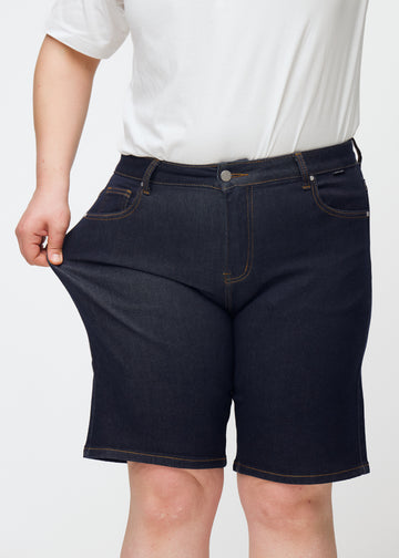 Plus-size model strækker dyb mørkeblå denim shorts til kvinder i farven Midnights ved låret for at vise materialets stretch.