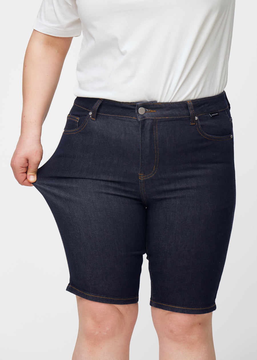Plus-size model strækker dyb mørkeblå denim shorts til kvinder i farven Midnights ved låret for at vise materialets stretch.