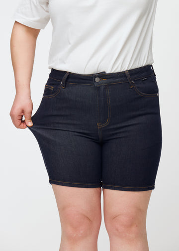 Plus-size model strækker dyb mørkeblå denim shorts til kvinder i farven Midnights ved låret for at vise materialets stretch.