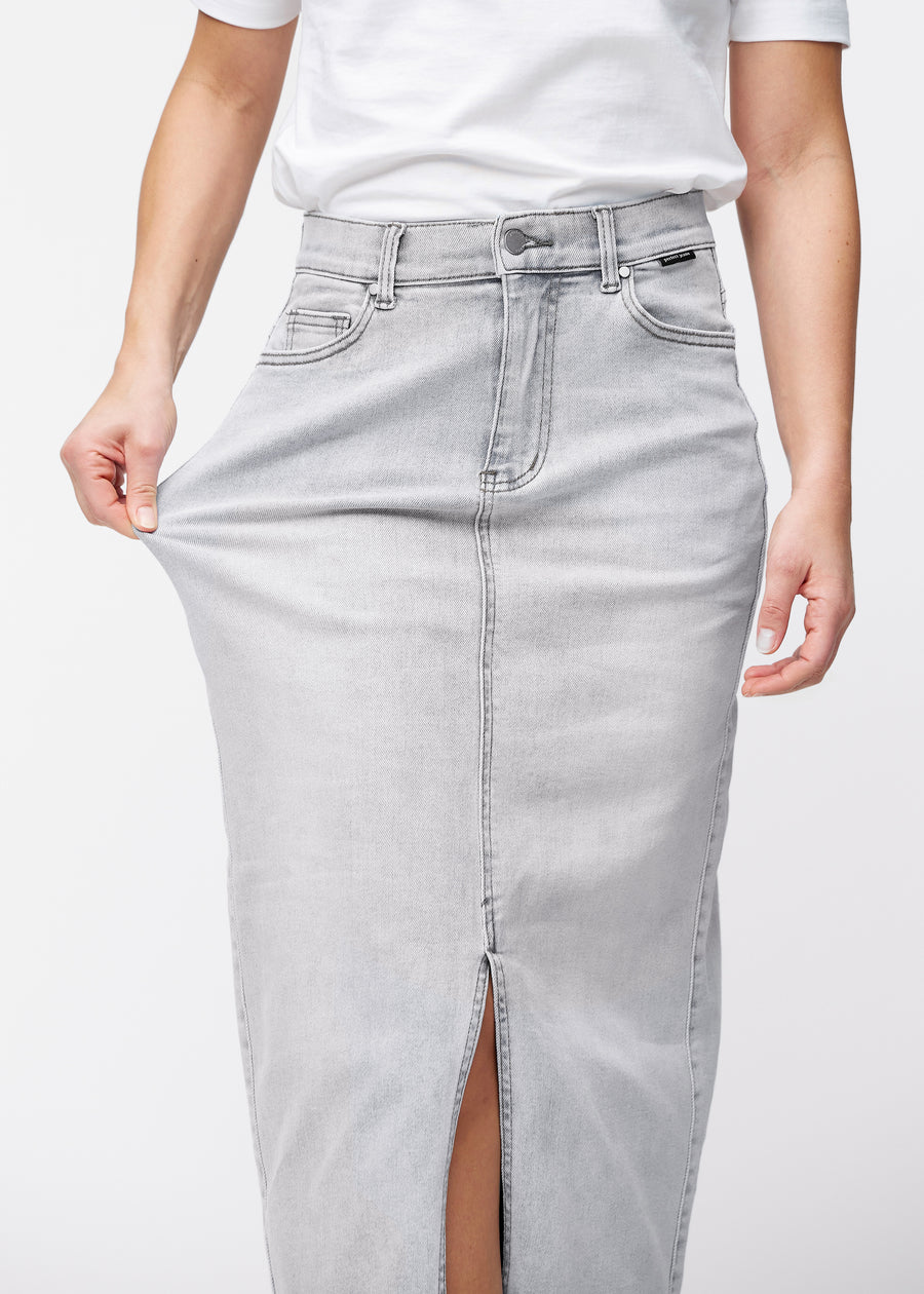 Model strækker den lange, lysegrå denimnederdel i farven Ashes ved hoften for at vise materialets stretch.