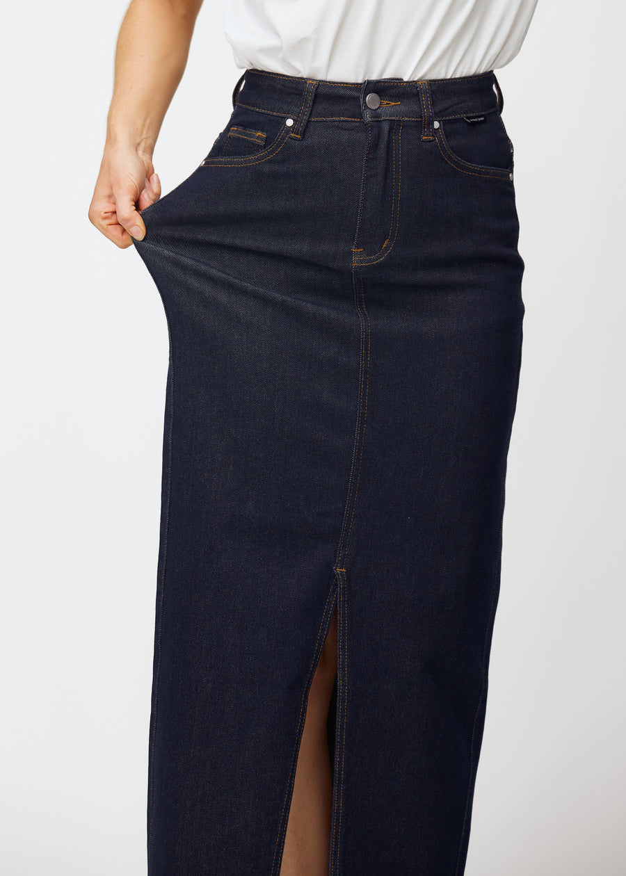 Model strækker den lange dyb mørkeblå denim nederdel til kvinder i farven Midnights ved hoften for at vise materialets stretch.