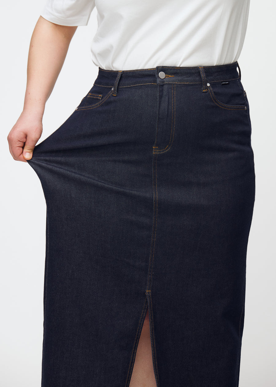 Plus-size model strækker den lange dyb mørkeblå denim nederdel til kvinder i farven Midnights ved hoften for at vise materialets stretch.