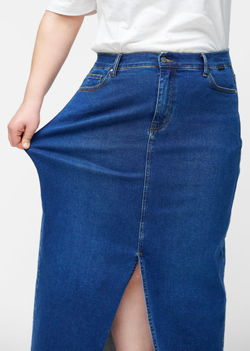 Plus-size model strækker den lange, mørkeblå denimnederdel i farven Oceans ved hoften for at vise materialets stretch.