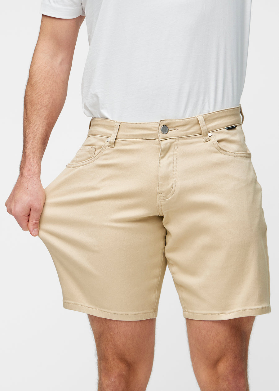 Model strækker beige shorts til mænd i farven Gazelles ved låret for at vise materialets stretch.