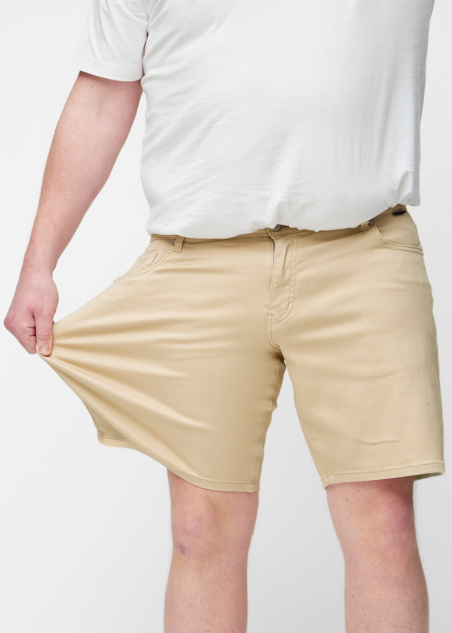 Plus-size model strækker beige shorts til mænd i farven Gazelles ved låret for at vise materialets stretch.