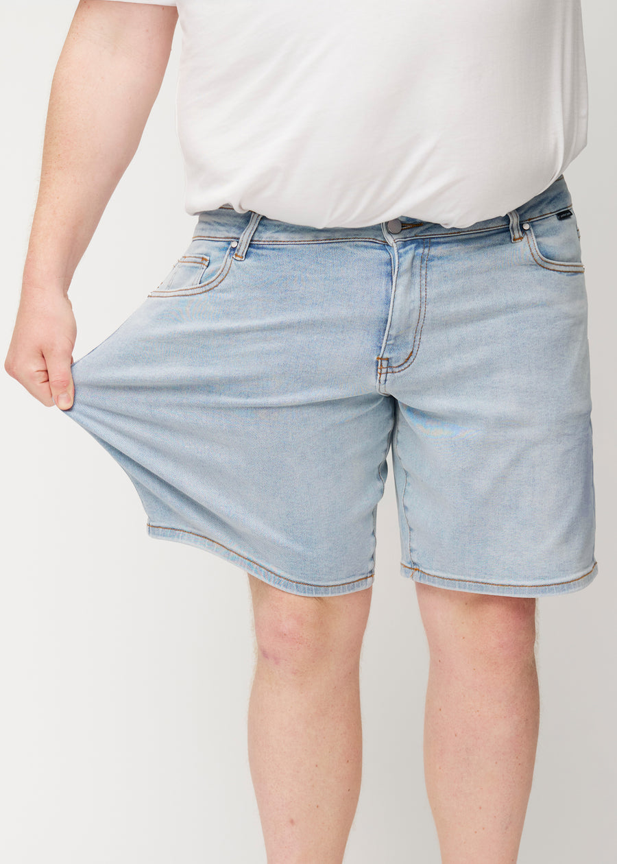 Plus-size model strækker lyseblå denim shorts til mænd i farven Waves ved låret for at vise materialets stretch.
