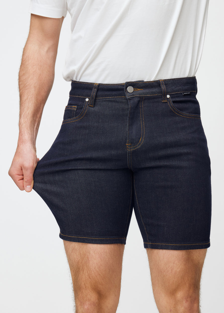 Model strækker dyb mørkeblå denim shorts til mænd i farven Midnights ved låret for at vise materialets stretch.