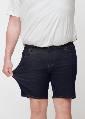 Plus-size model strækker dyb mørkeblå denim shorts til mænd i farven Midnights ved låret for at vise materialets stretch.