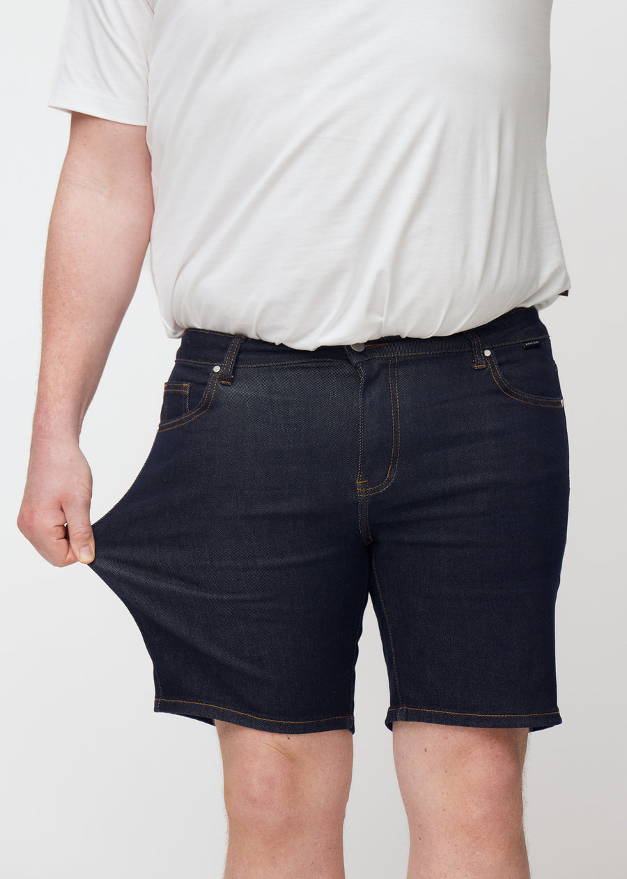 Plus-size model strækker dyb mørkeblå denim shorts til mænd i farven Midnights ved låret for at vise materialets stretch.